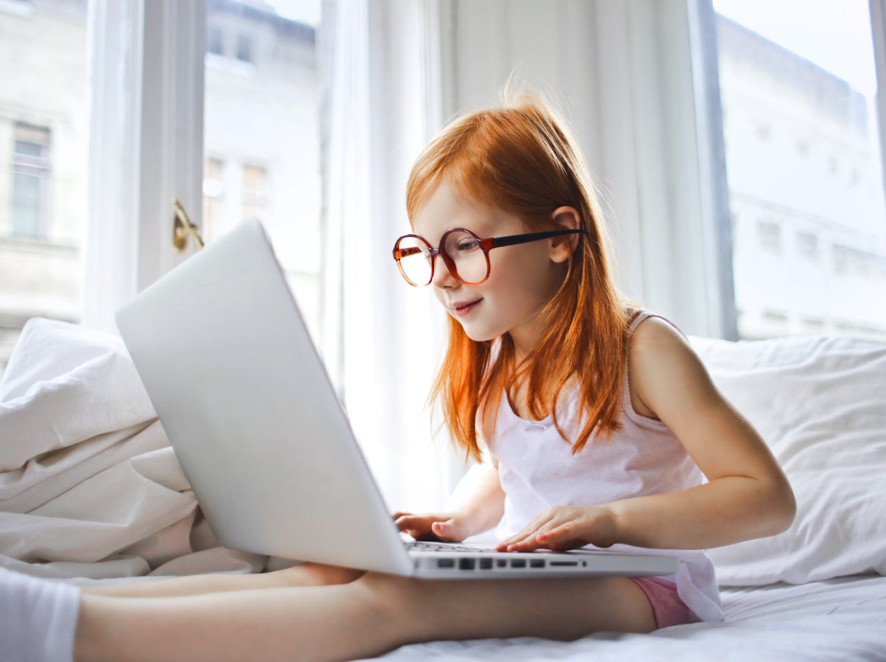 Red-haired child in glasses using a laptop on a white bed, bright room.